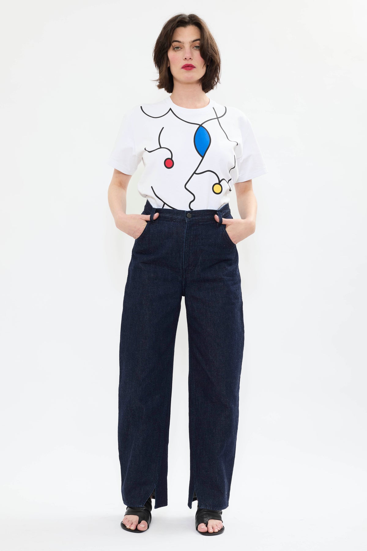 Model wearing AYT STUDIO 'TRIBE' high-rise curved straight leg 100% recycled cotton jeans in blue with signature AYT Studio logo, ankle slit detail, and functional front and back pockets.