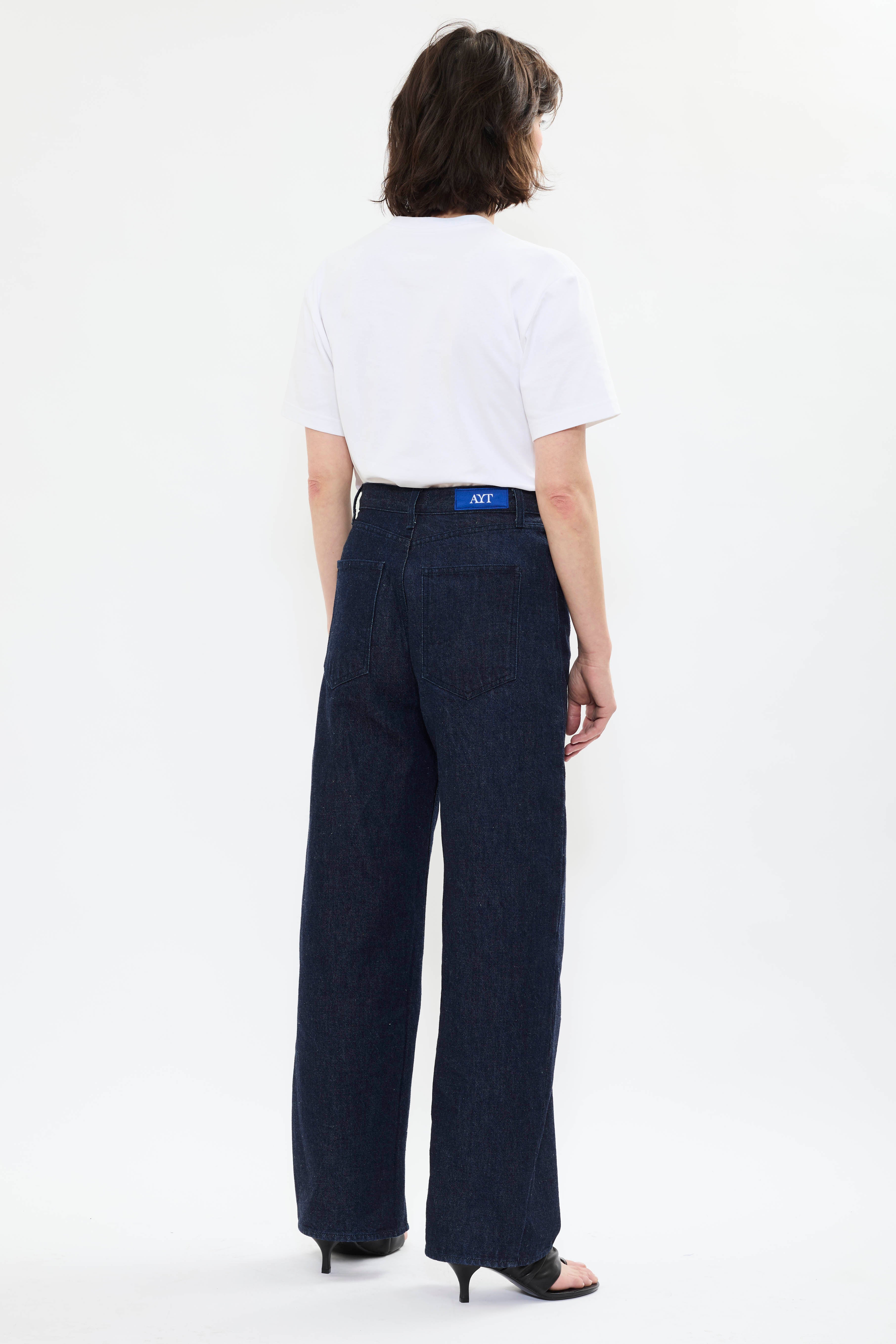 Back of AYT STUDIO 'TRIBE' high-rise curved straight leg 100% recycled cotton jeans in blue with signature AYT Studio logo, ankle slit detail, and functional front and back pockets.