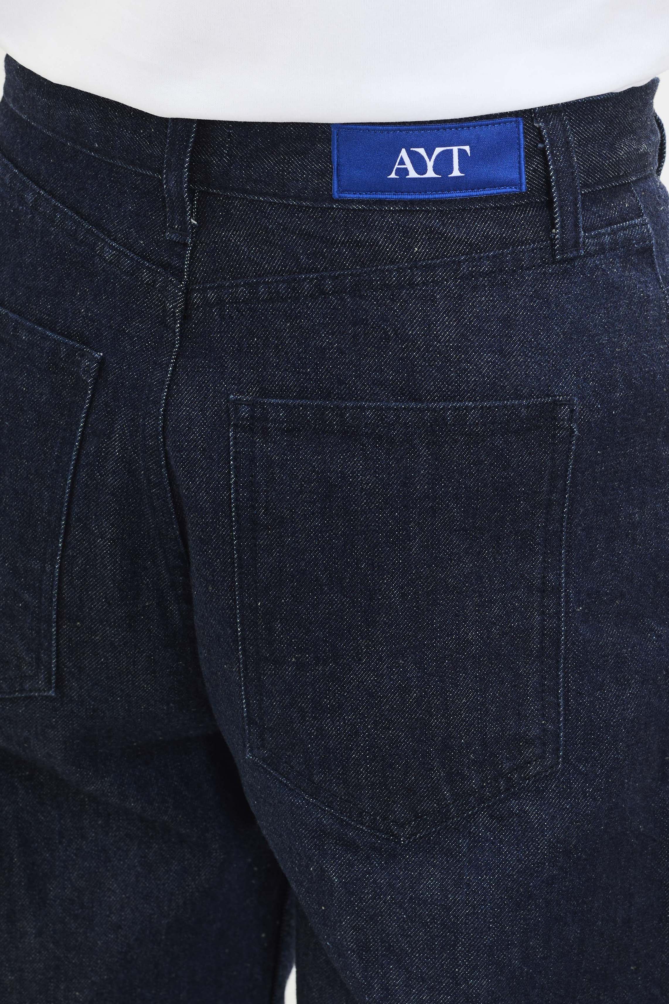 Details of AYT STUDIO 'TRIBE' high-rise curved straight leg 100% recycled cotton jeans in blue with signature AYT Studio logo, ankle slit detail, and functional front and back pockets.