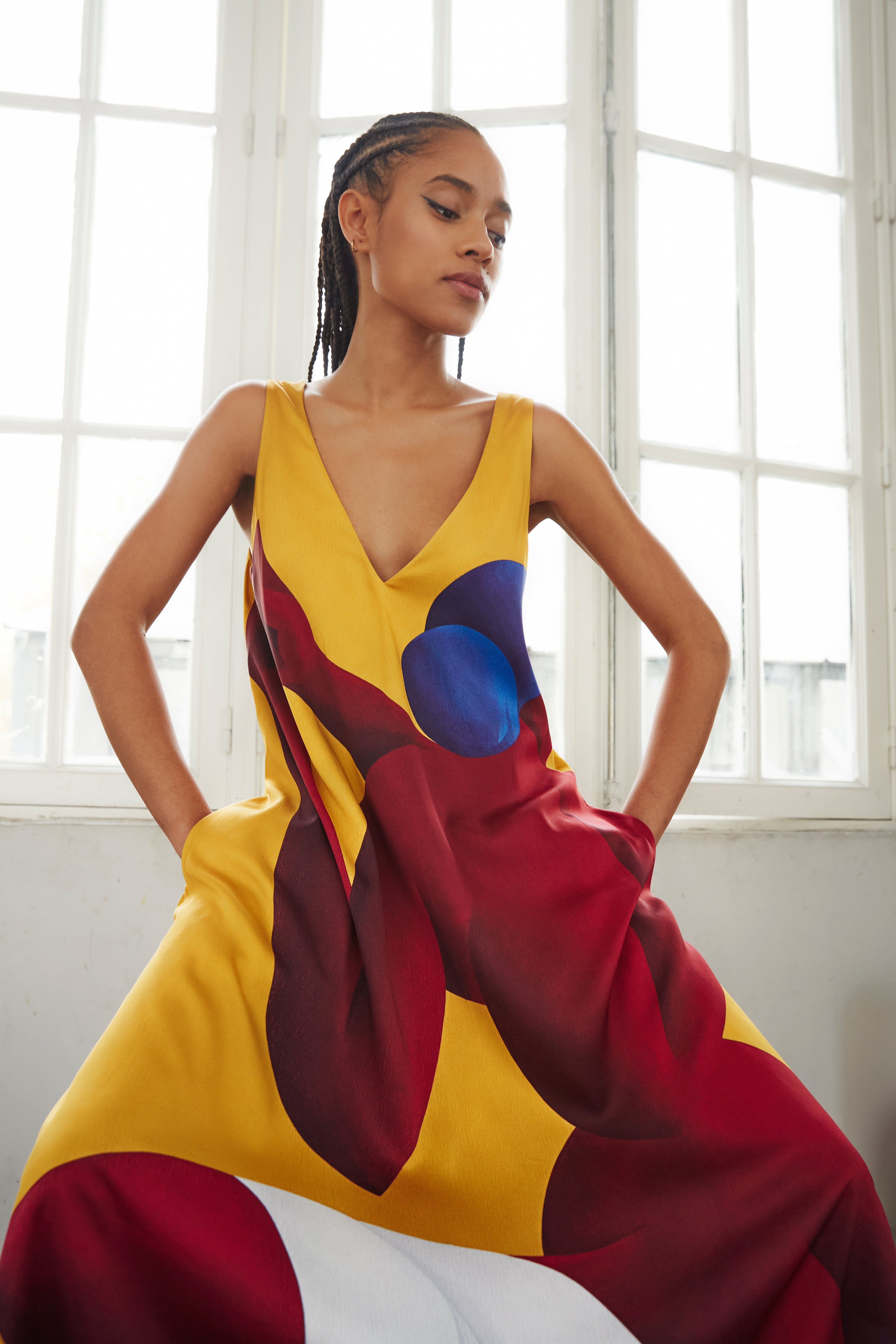 Woman modeling AYT Studio x French artist REMED 'SORORITY' fluid, vibrant yellow printed dress in viscose and elastane with punging V-neck front and back and discreet side pockets. Inspired by REMED's 'Bain de sable.'