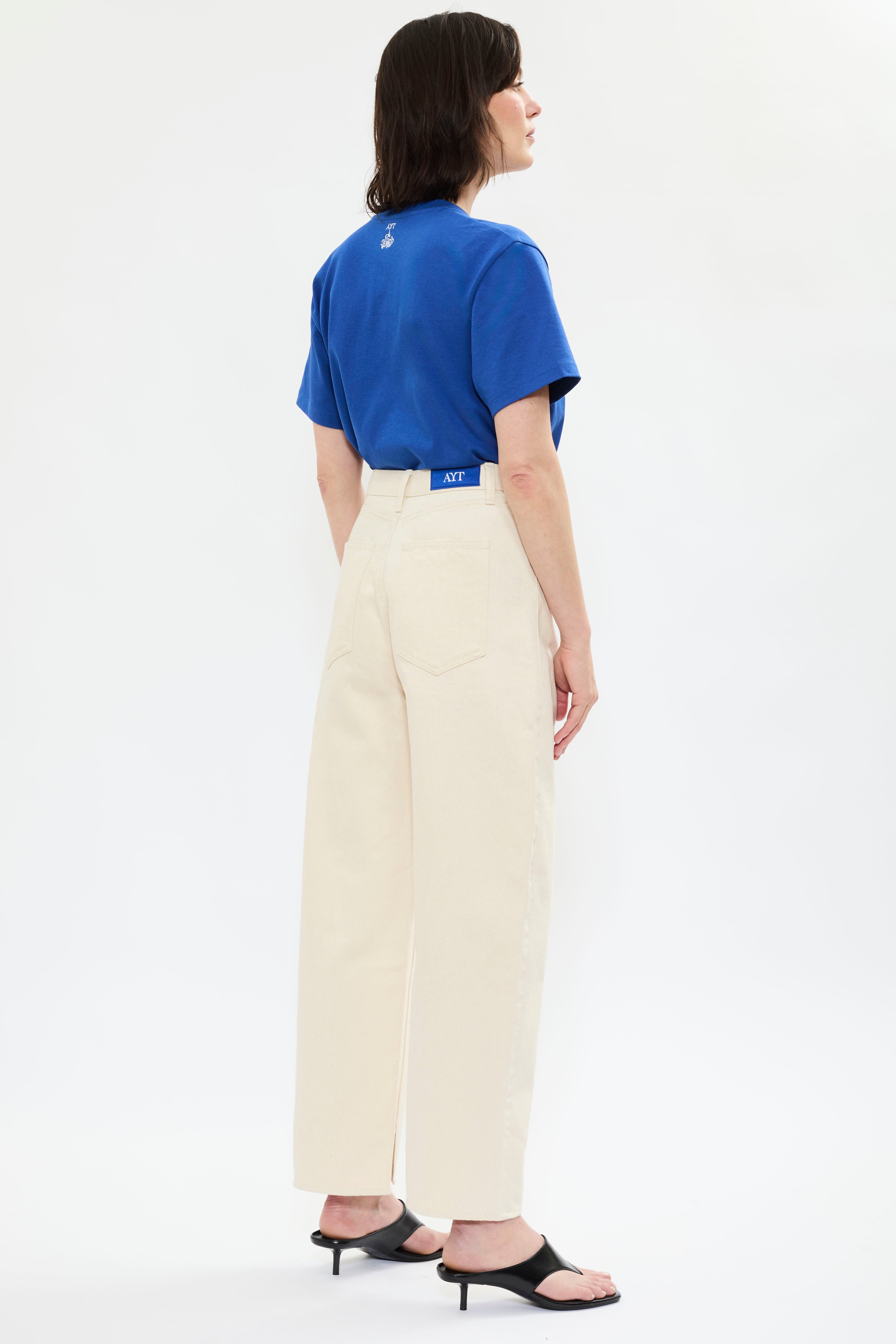 Rear of AYT STUDIO 'TRIBE' high-rise curved straight leg 100% recycled cotton jeans in cream with signature blue AYT Studio logo, ankle slit detail, and functional front and back pockets.