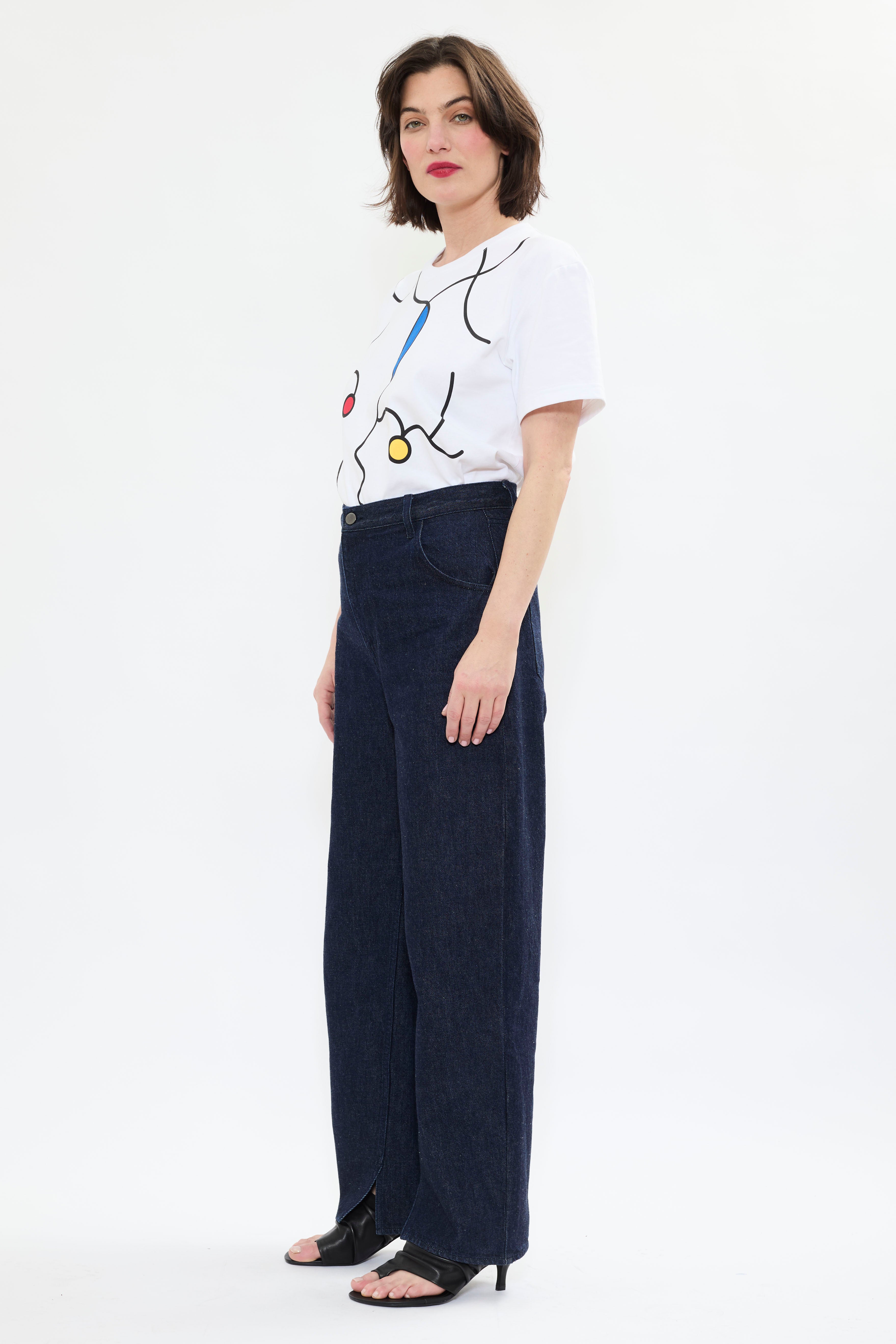Profile of model in AYT STUDIO 'TRIBE' high-rise curved straight leg 100% recycled cotton jeans in blue with signature AYT Studio logo, ankle slit detail, and functional front and back pockets.