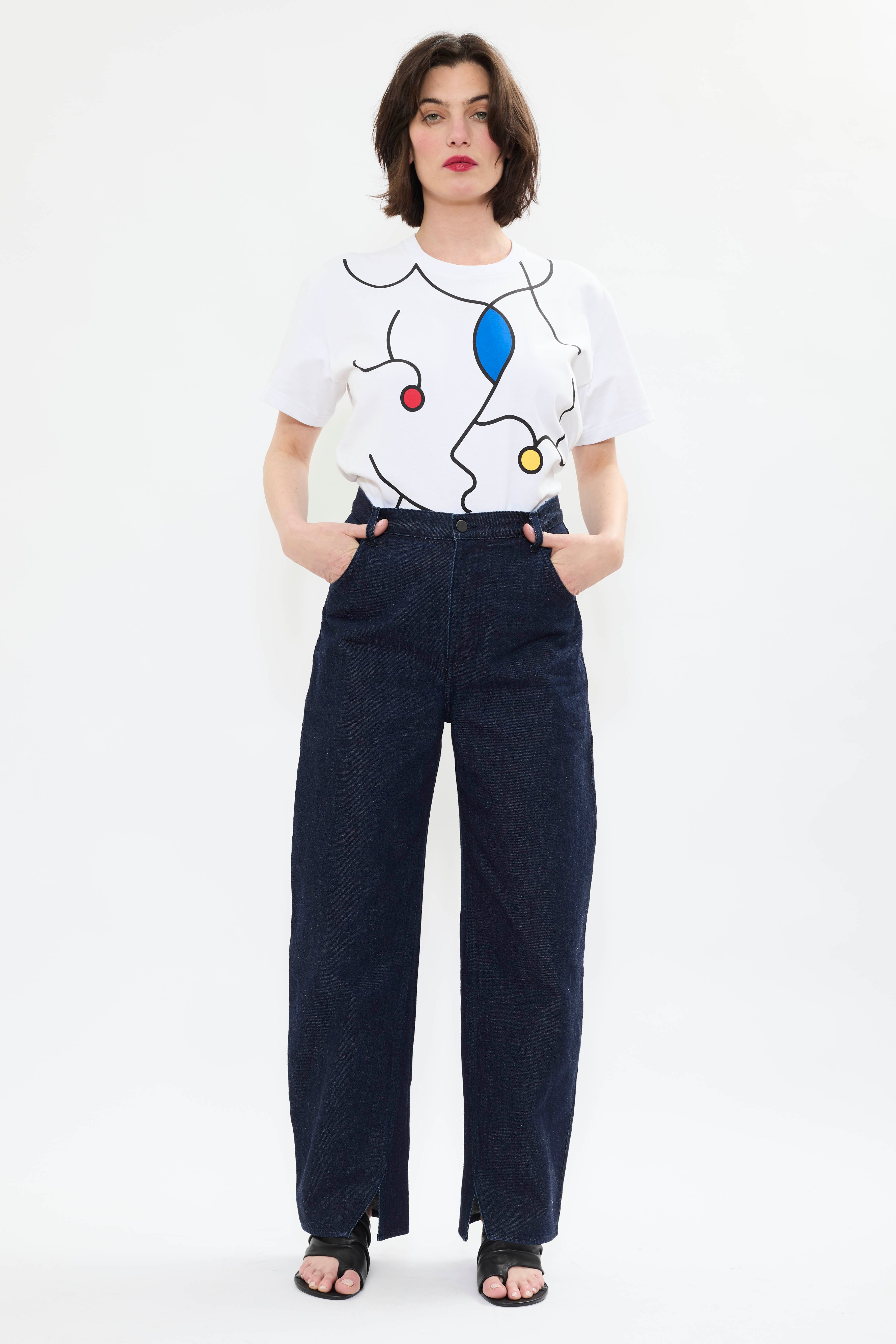 Model wearing AYT STUDIO 'TRIBE' high-rise curved straight leg 100% recycled cotton jeans in blue with signature AYT Studio logo, ankle slit detail, and functional front and back pockets.