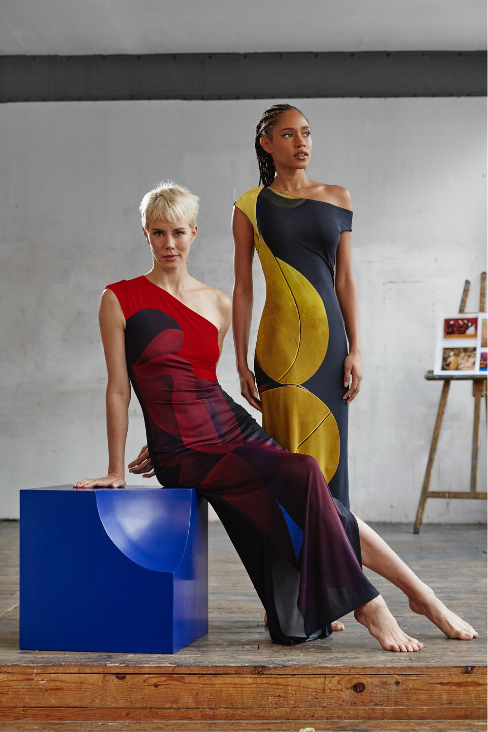 Models wearing AYT STUDIO x REMED, 'AURA' fluid viscose and elastane maxi dress with asymmetrical shoulder and side slits in printed black and gold, inspired by 'Femme & sphère.' Paired with 'MUSE' by AYT STUDIO x REMED.