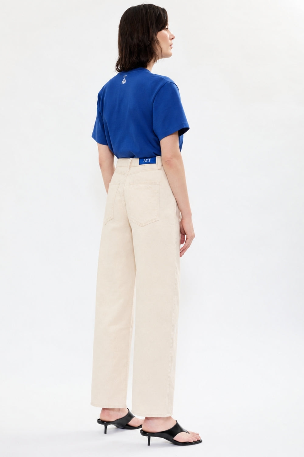 Back of AYT STUDIO 'TRIBE' high-rise curved straight leg 100% recycled cotton jeans in cream with signature blue AYT Studio logo, ankle slit detail, and functional front and back pockets.
