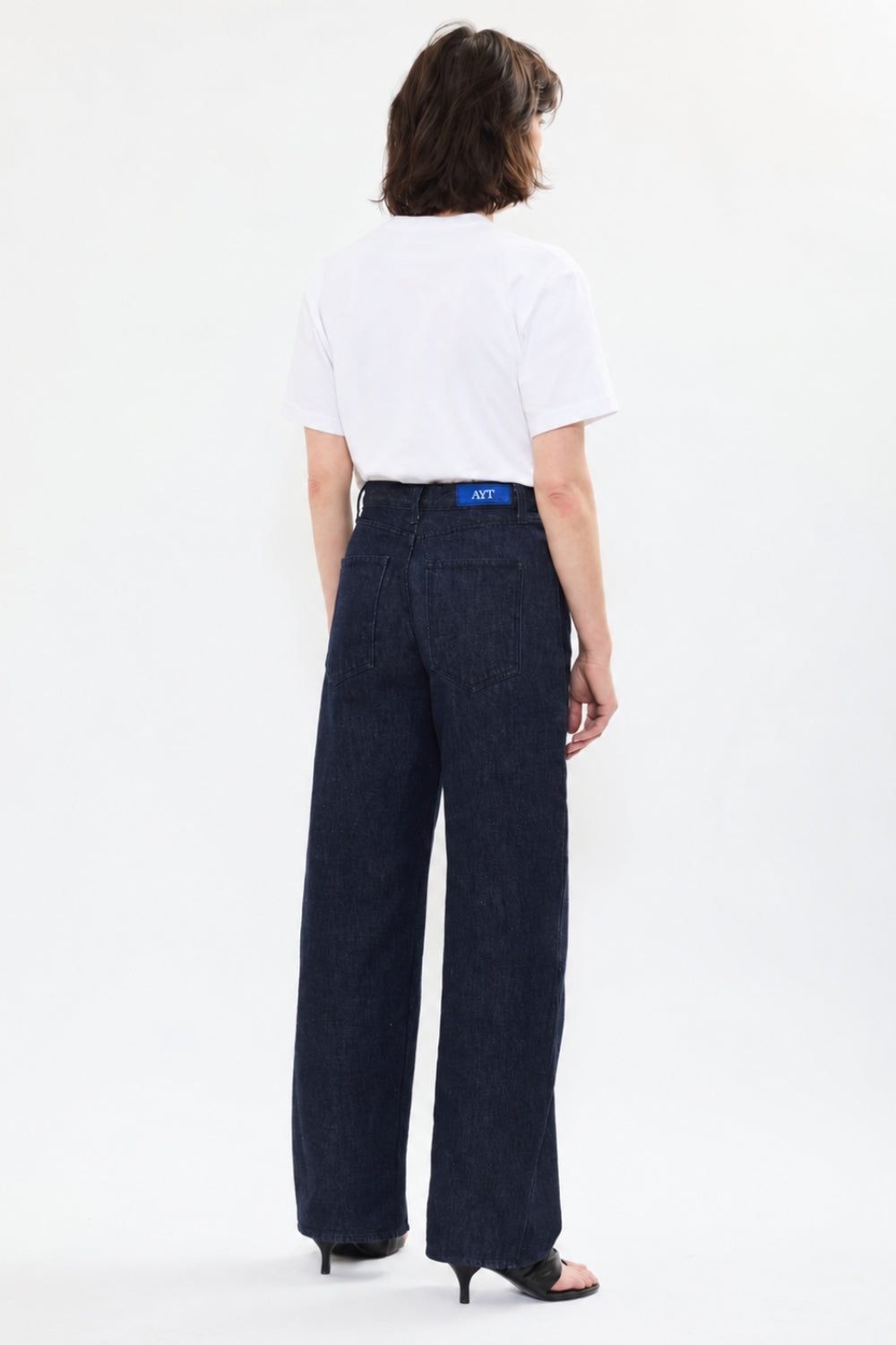 Back of AYT STUDIO 'TRIBE' high-rise curved straight leg 100% recycled cotton jeans in blue with signature AYT Studio logo, ankle slit detail, and functional front and back pockets.